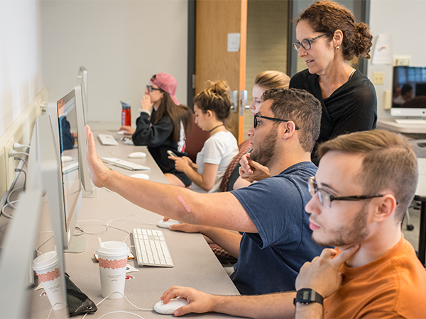 A female instructor stands over a group of students reviewing content on a row of computers. One of the students is pointing out information on the screen to the instructor.